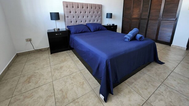 Un dormitorio con cabecera capitonada, cama con ropa de cama azul, dos mesitas de noche con lámparas y un armario con puertas correderas.