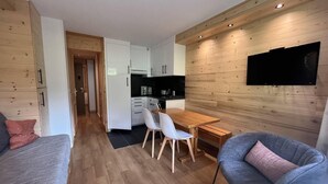 A modern apartment with a wooden floor, a dining area with a table and chairs, a kitchen with white cabinets, and a flat-screen TV mounted on the wall. - 20m from ski slopes, 1st floor, view ski slopes, terrace, ski locker, 27m², Méribel-Mottaret (Méribel - Les Allues)