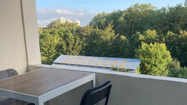 En balkong med ett bord och stolar, med utsikt över en stadsmiljö med träd och byggnader.