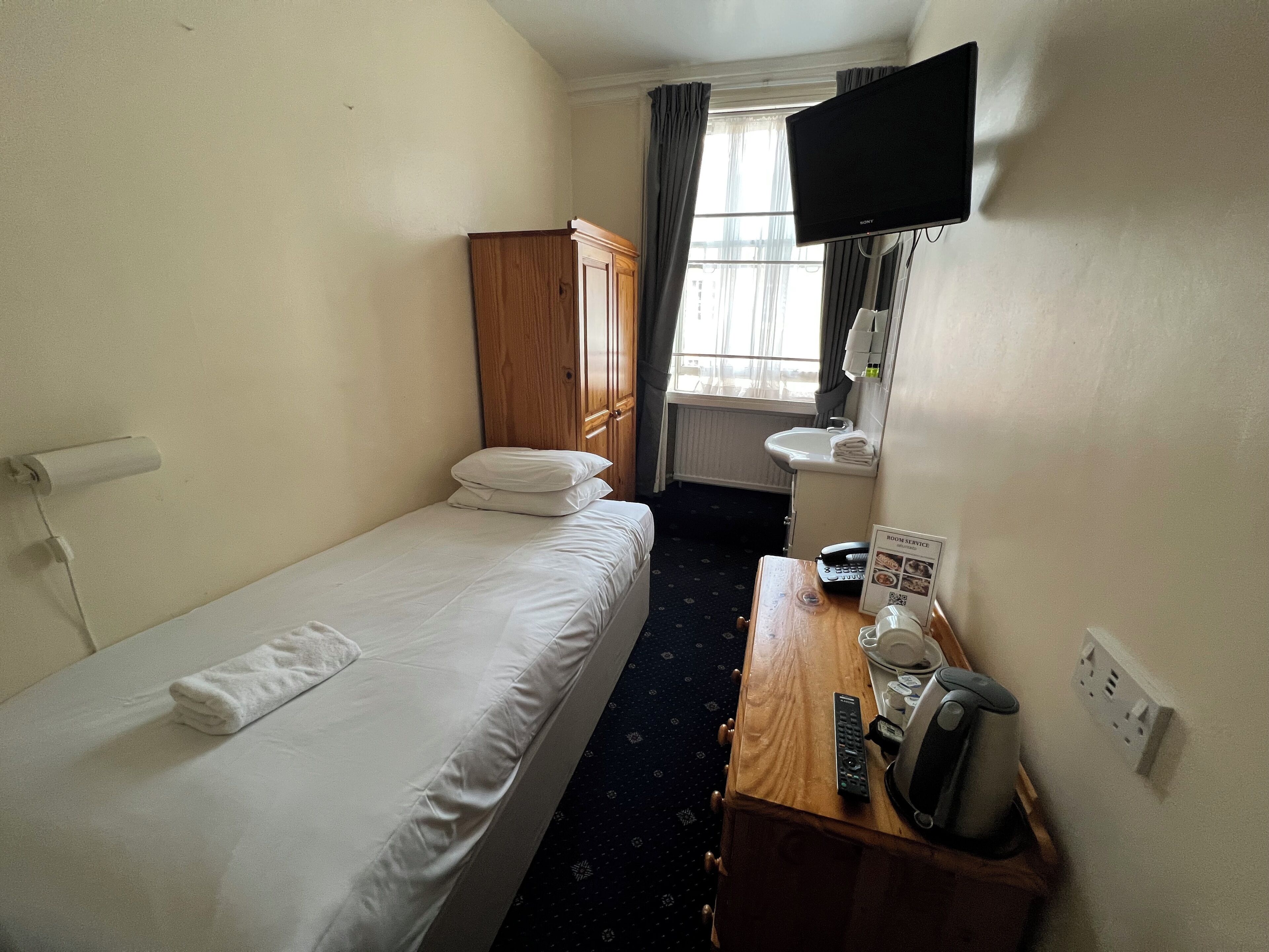 A hotel room with a single bed, a wooden wardrobe, a small desk with a telephone and a coffee maker, a TV mounted on the wall, and a window with curtains.