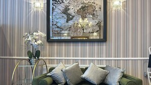 Een groene bank met meerdere kussens, een klein tafeltje met een vaas witte bloemen, en een wereldkaart in een lijst aan de muur.