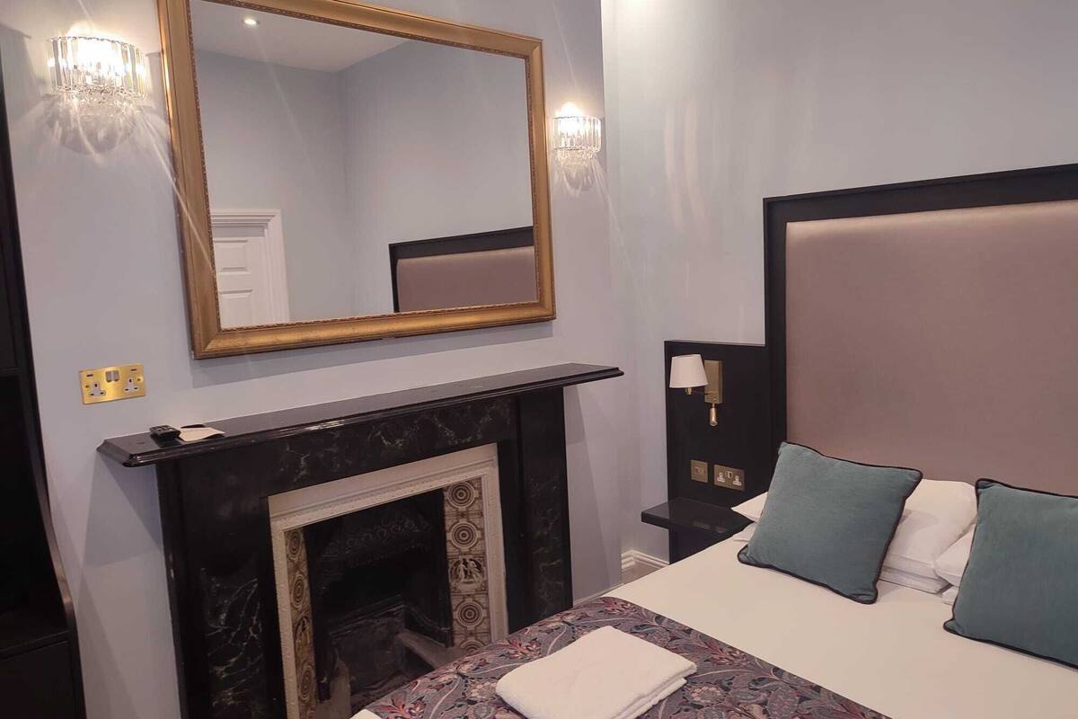 A hotel room with a large bed, a fireplace, a mirror, and a nightstand with a lamp.