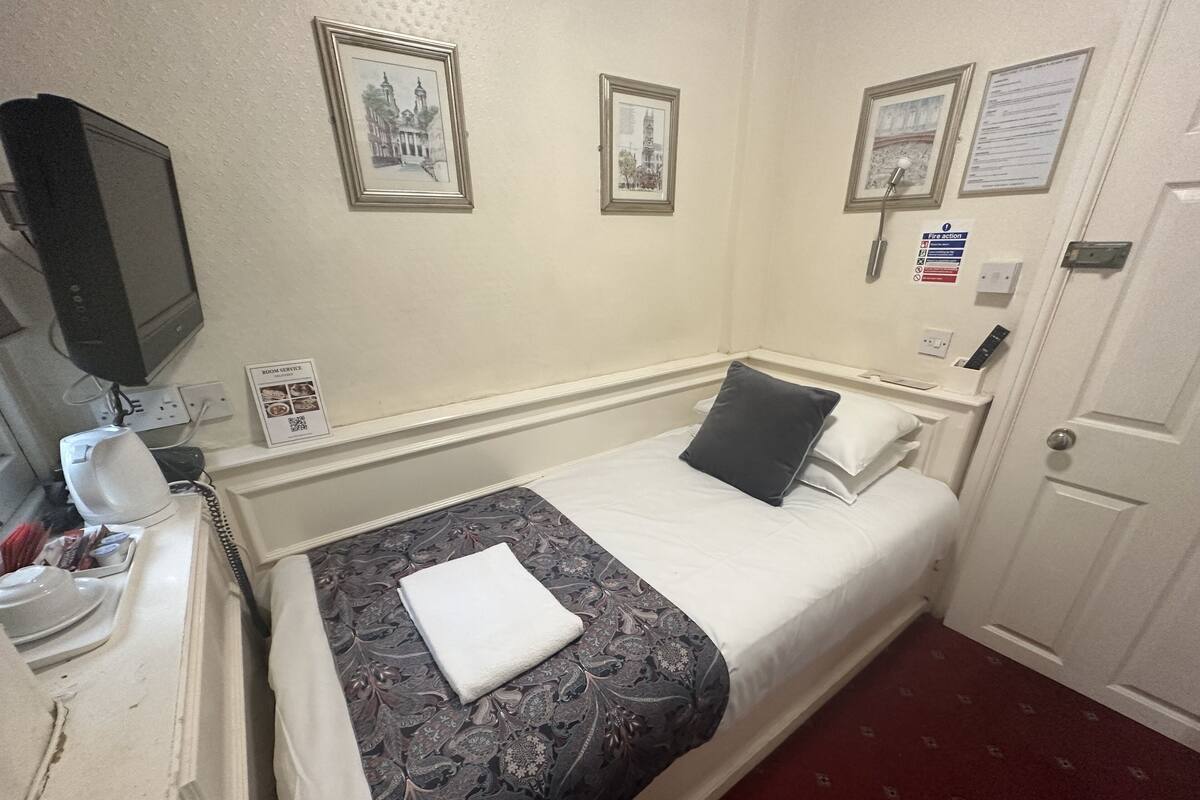 A hotel room with a bed, a bedside table with a lamp, a telephone, and framed pictures on the wall.