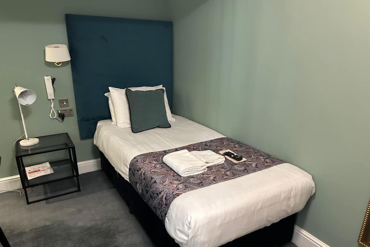 A neatly made bed with a patterned blanket, a bedside lamp, and a small table with a lamp and a phone.