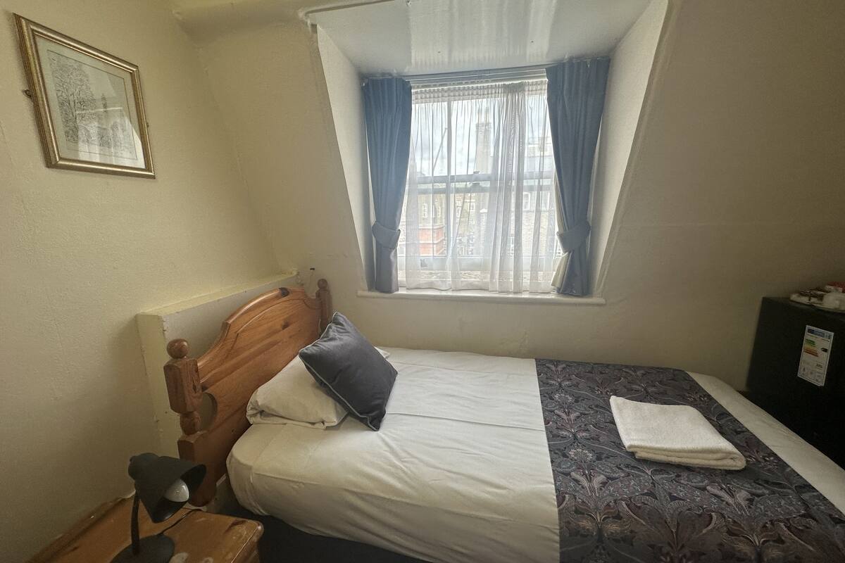 A small, simple room with a single bed, a wooden headboard, a desk with a lamp, and a window with curtains.