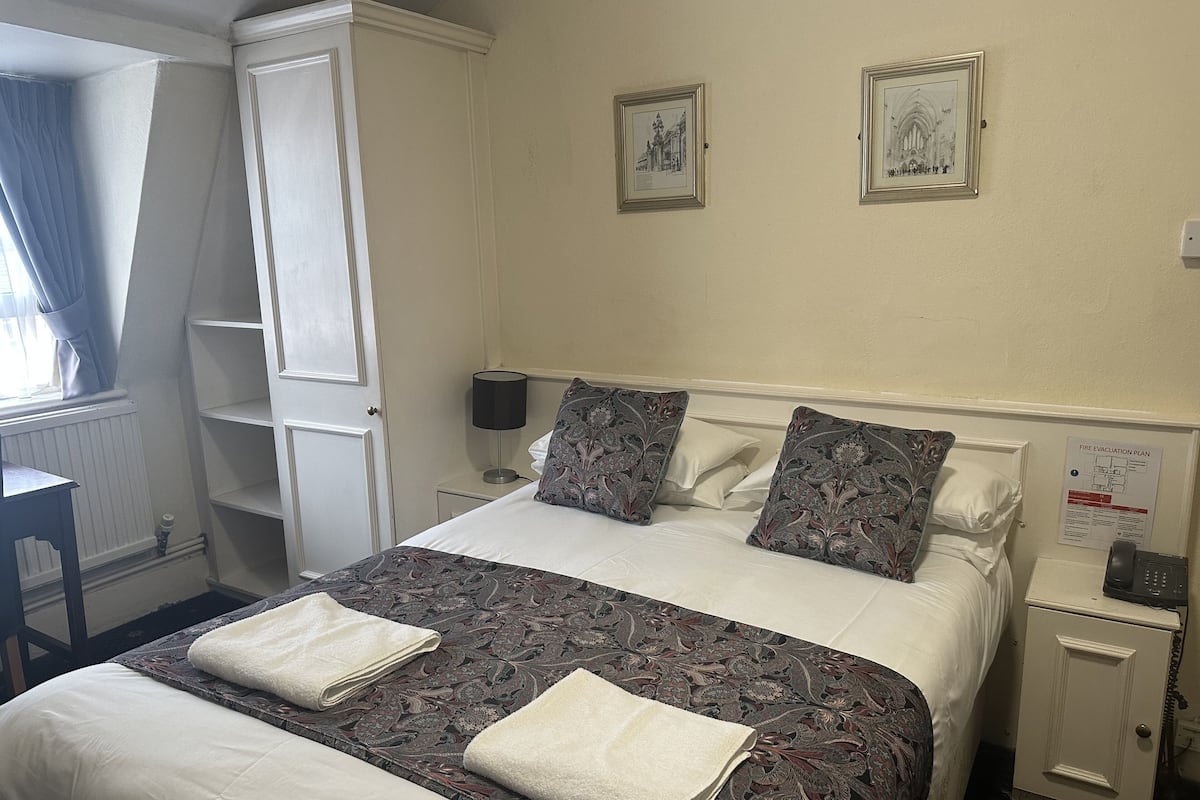 A neatly made bed with patterned bedding and two pillows, a bedside table with a lamp, and a wall-mounted fire evacuation plan.