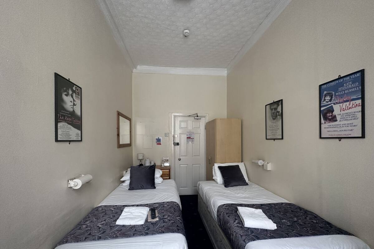 A hotel room with two single beds, a door, a mirror, a poster, and a small table with a lamp.