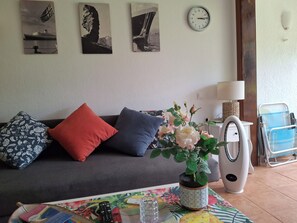 Una sala de estar con un sofá, almohadas, un reloj, una planta, una lámpara, un ventilador y un control remoto sobre una mesa.