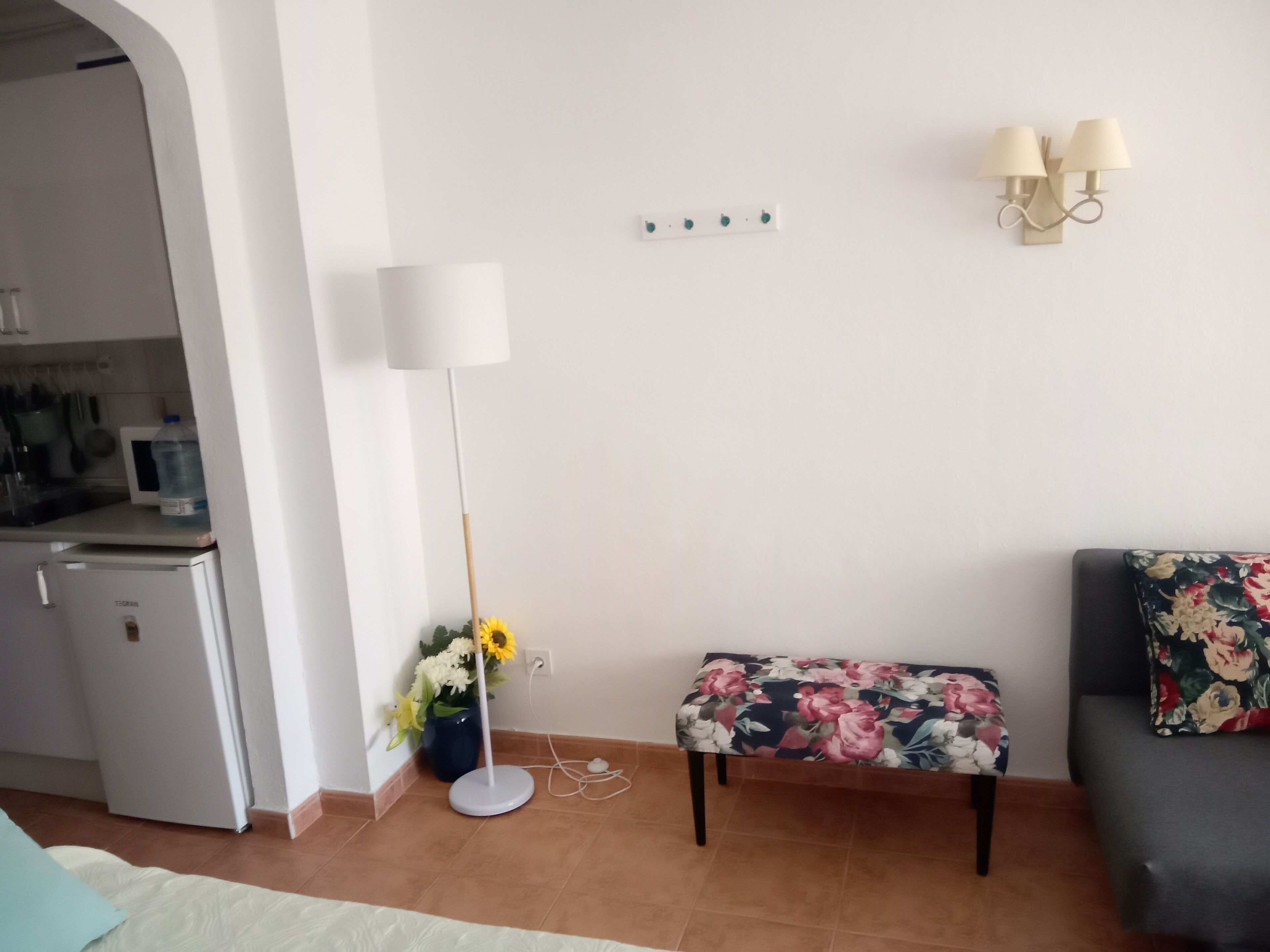 Een knus woongedeelte met een kitchenette, een staande vloerlamp, een bankje met bloemenprint en een bank met een sierkussen met bloemenmotief.