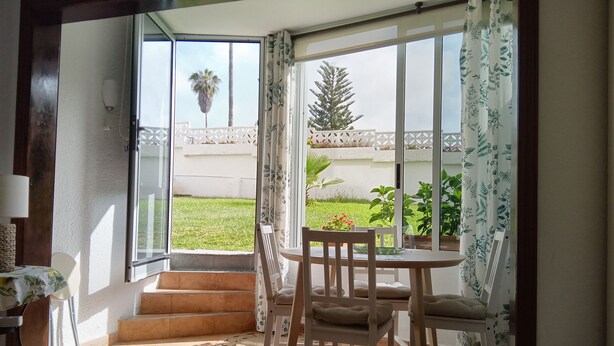 A room with a sliding glass door leading to a balcony with a view of a lawn and a palm tree. There is a small table with chairs and a potted plant on the balcony.