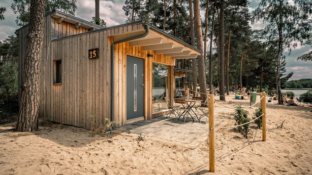 A wooden cabin with a number '15' on its door, situated on a sandy beach with trees in the background. - Naturcamping Brettmühlenteich e.G. (Thiendorf)