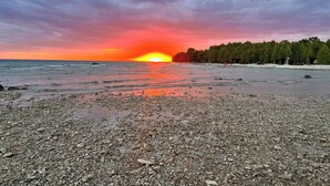 Uma praia de seixos com o pôr do sol no oceano, árvores ao fundo e uma bandeira em uma estrutura.