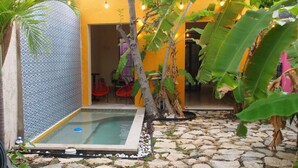 A small outdoor pool area with a stone floor, surrounded by tropical plants and a yellow wall. - Encantadora Suite, en la Mejor Zona de Paseo Montejo. En el Barrio Gastronómico (Mérida)