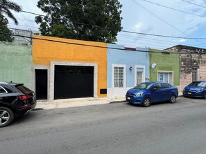 A street with buildings painted in various colors, a black car parked on the left, and a blue car parked on the right.
