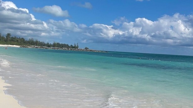 Une plage à l’eau turquoise et limpide, avec un rivage sablonneux et une côte lointaine parsemée de végétation et de rochers.