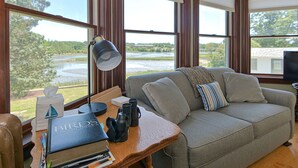 Ein gemütliches Zimmer mit einem grauen Sofa, einem Holztisch mit Büchern und einem Teleskop sowie großen Fenstern mit Blick auf einen Fluss und Grünflächen.