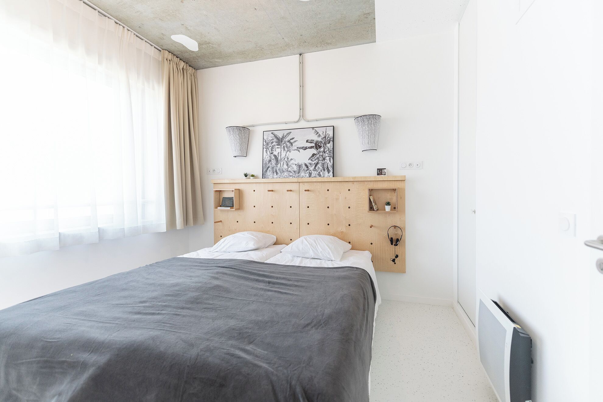 Un dormitorio minimalista con cama, cabecera de madera, estante fijado a la pared y ventana con cortinas.