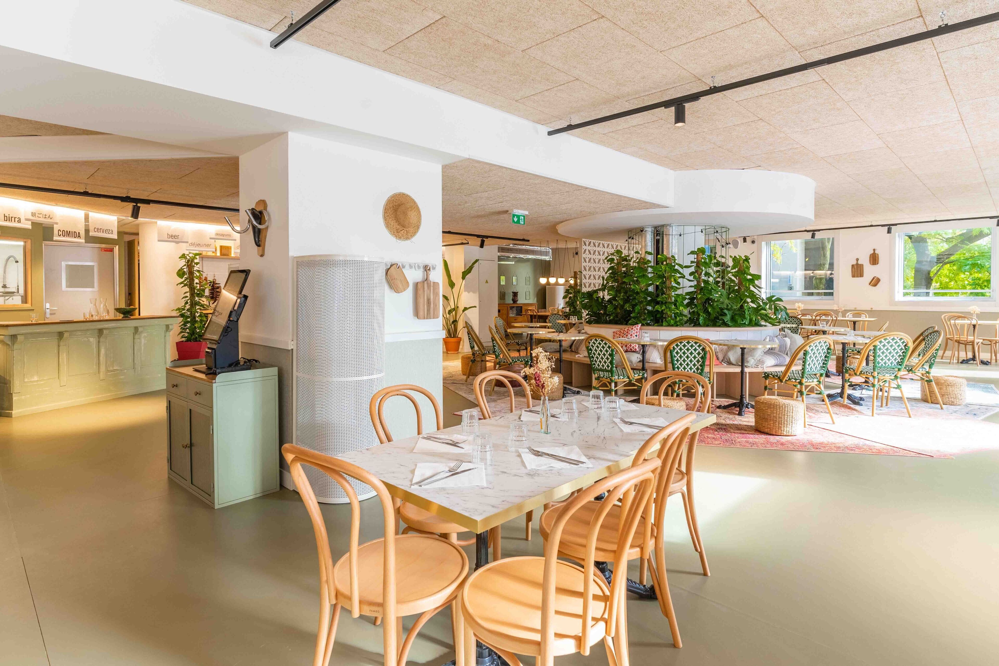 Un restaurante moderno con una zona central de comedor que incluye sillas de madera y una mesa de mármol. Hay plantas en macetas y una planta verde grande al fondo.