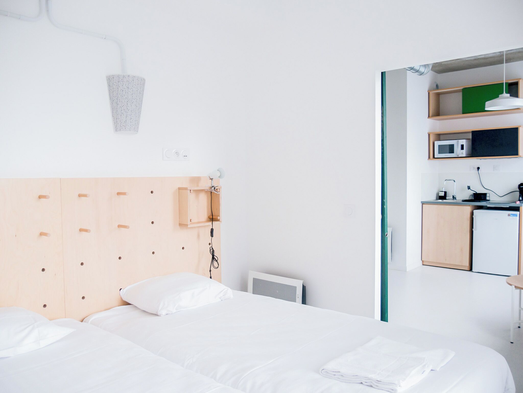 A modern hotel room with a wooden headboard, a bed with white linens, a small kitchenette with a microwave, and a wall-mounted light fixture.