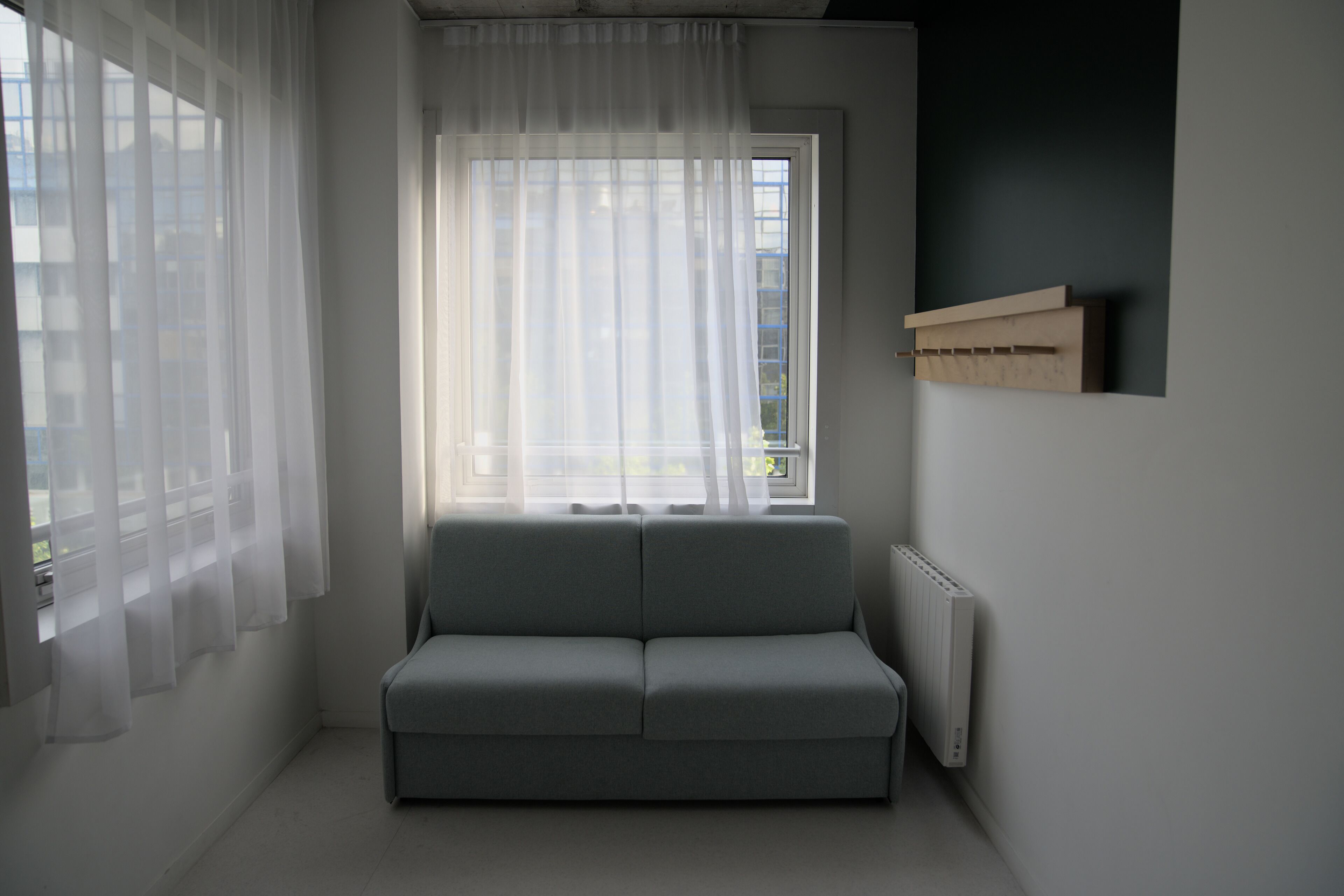 A modern living room with a grey sofa, sheer curtains, a radiator, and a window with a view of buildings.