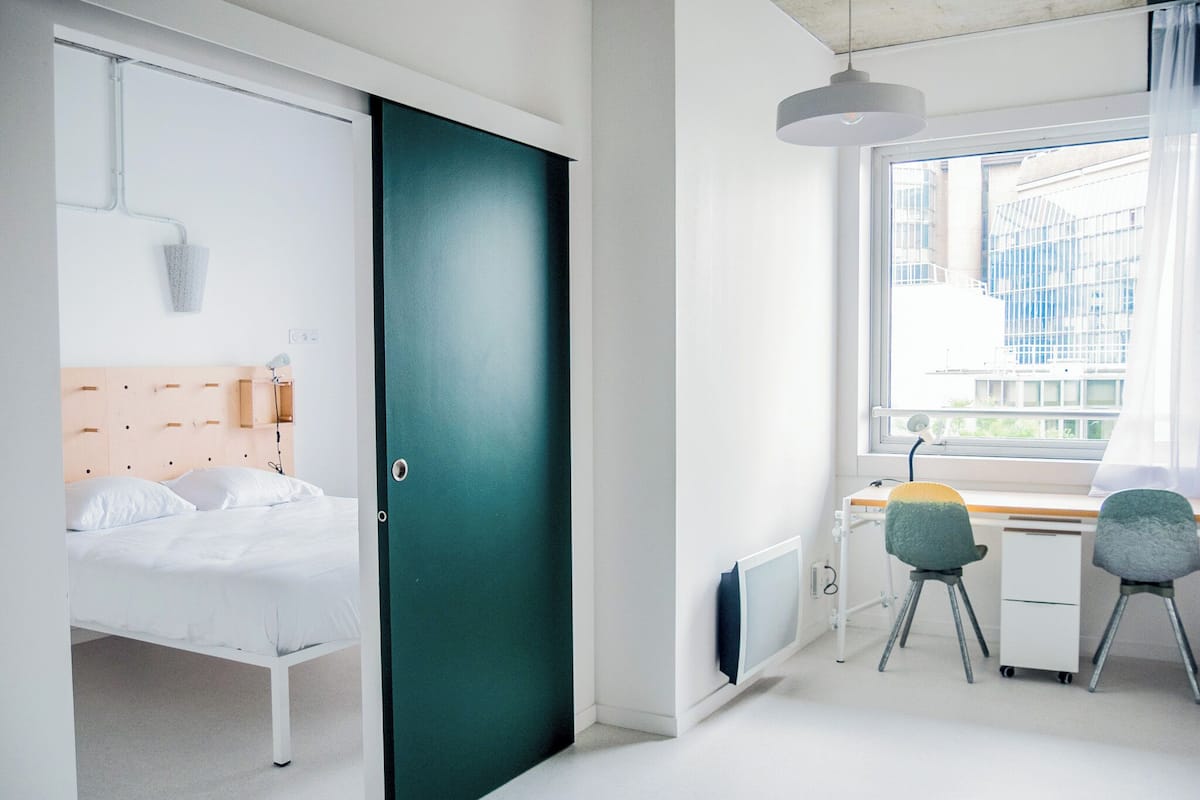 A modern bedroom with a bed, a desk, and a chair, a green door, and a window with a view of buildings.