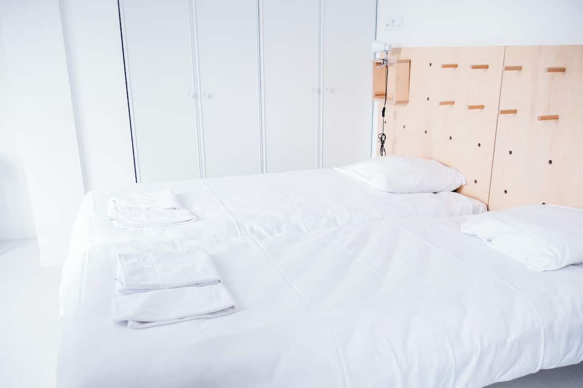 A neatly made bed with white linens and pillows, a wooden headboard with a light switch, and a small wooden shelf with a power outlet.