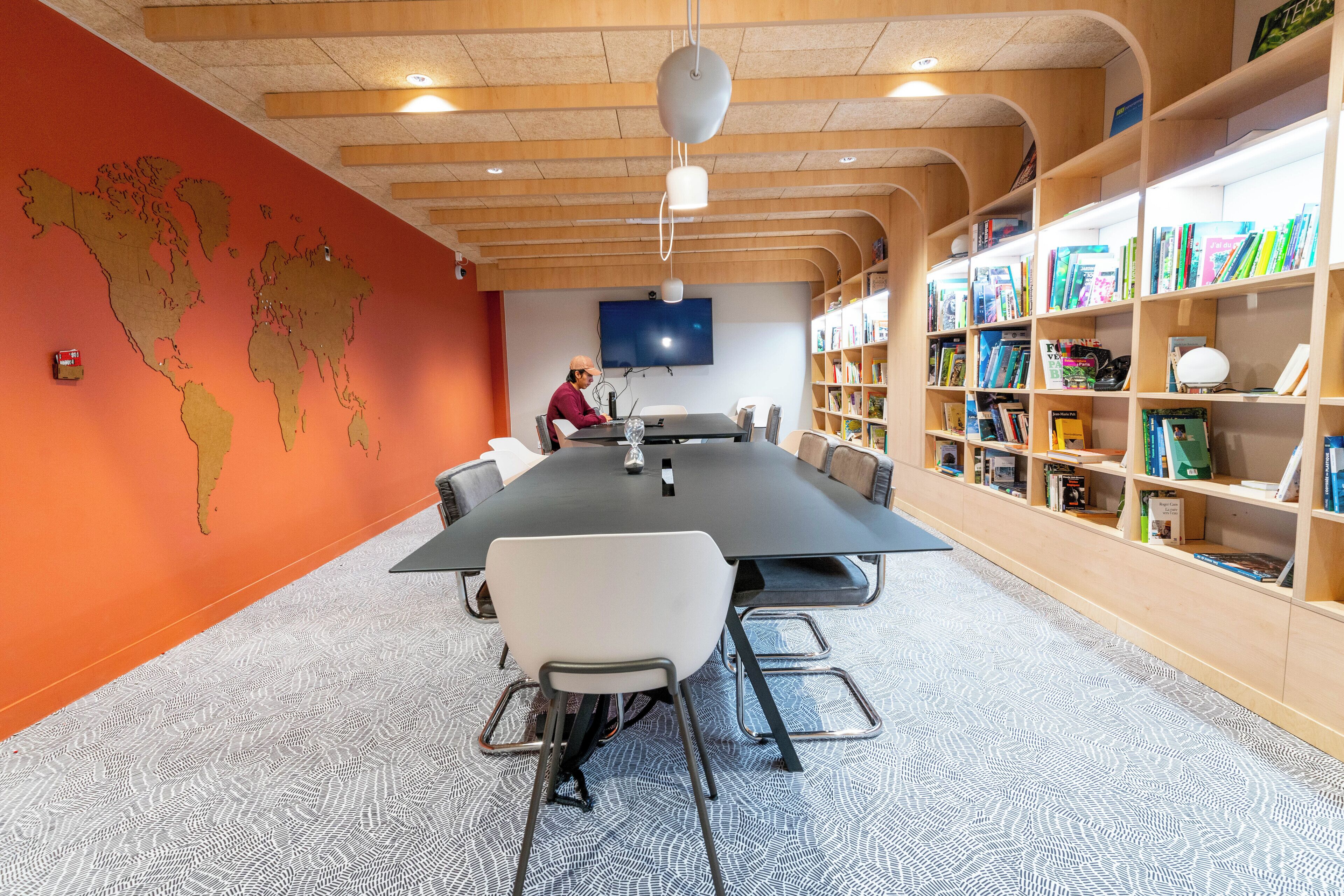 Een vergaderruimte met een grote tafel, stoelen, een wereldkaart aan de muur, een televisie en boekenkasten.