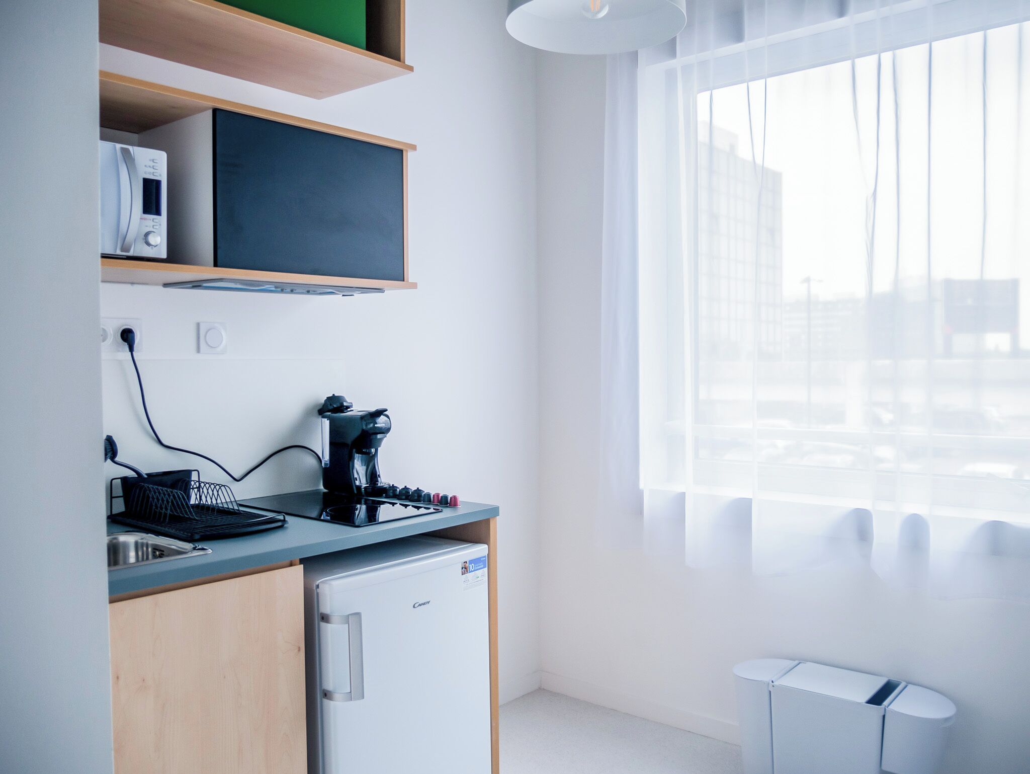 A modern kitchen with a coffee maker, a small refrigerator, and a window with sheer curtains.