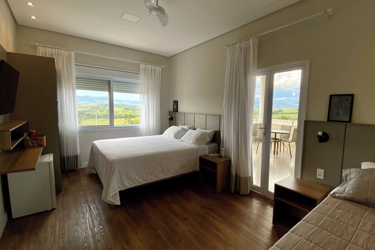 A bedroom with a large bed, a smaller bed, a desk, and a chair. There is a window with a view of the countryside.