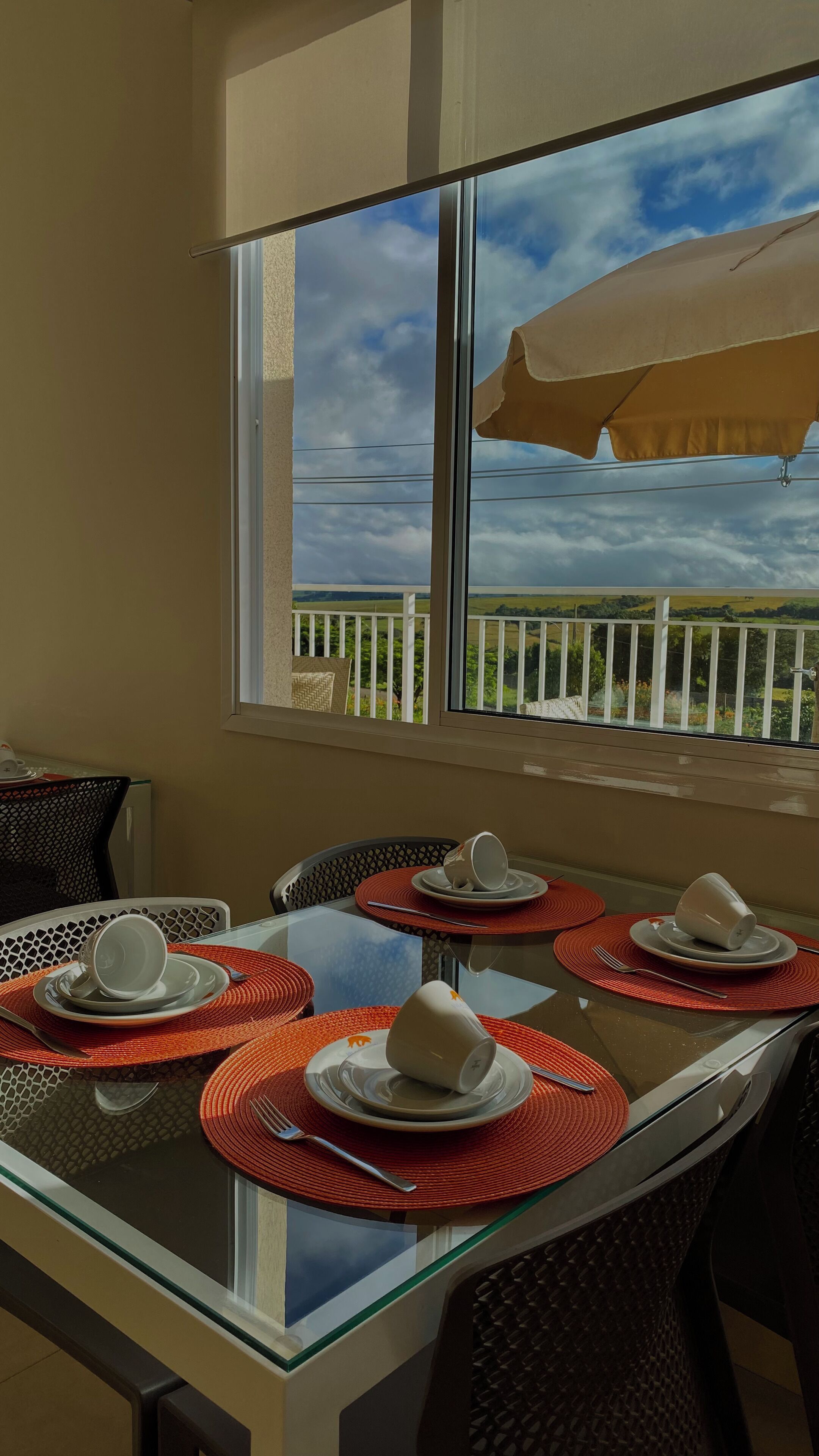Uma área de refeições com mesa de vidro, jogos americanos vermelhos e pratos e copos brancos.