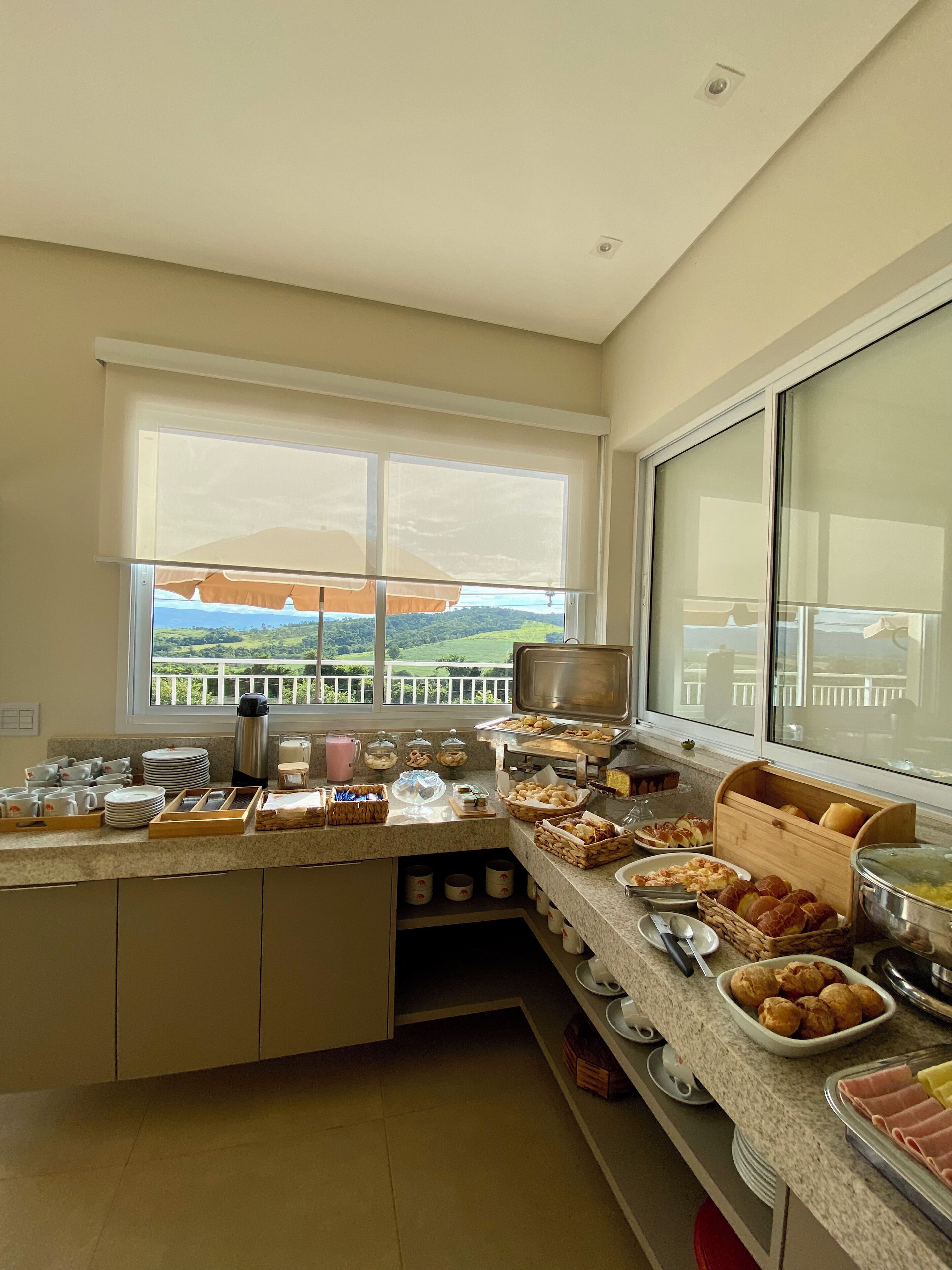 Cozinha com disposição de buffet, uma janela grande com vista para o verde e diversos itens alimentares à mostra.
