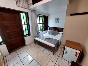Un dormitorio con cama, un banco de madera, un radiador, una ventana con cortinas y un refrigerador pequeño.