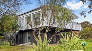 Une maison moderne à deux étages, dotée d’une façade en verre, d’une terrasse en bois et d’un escalier menant à l’entrée.