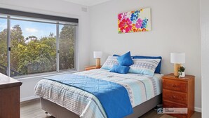 Une chambre avec une grande fenêtre, un lit avec une literie bleue, deux tables de chevet avec des lampes et une œuvre d’art florale aux couleurs vives accrochée au mur.