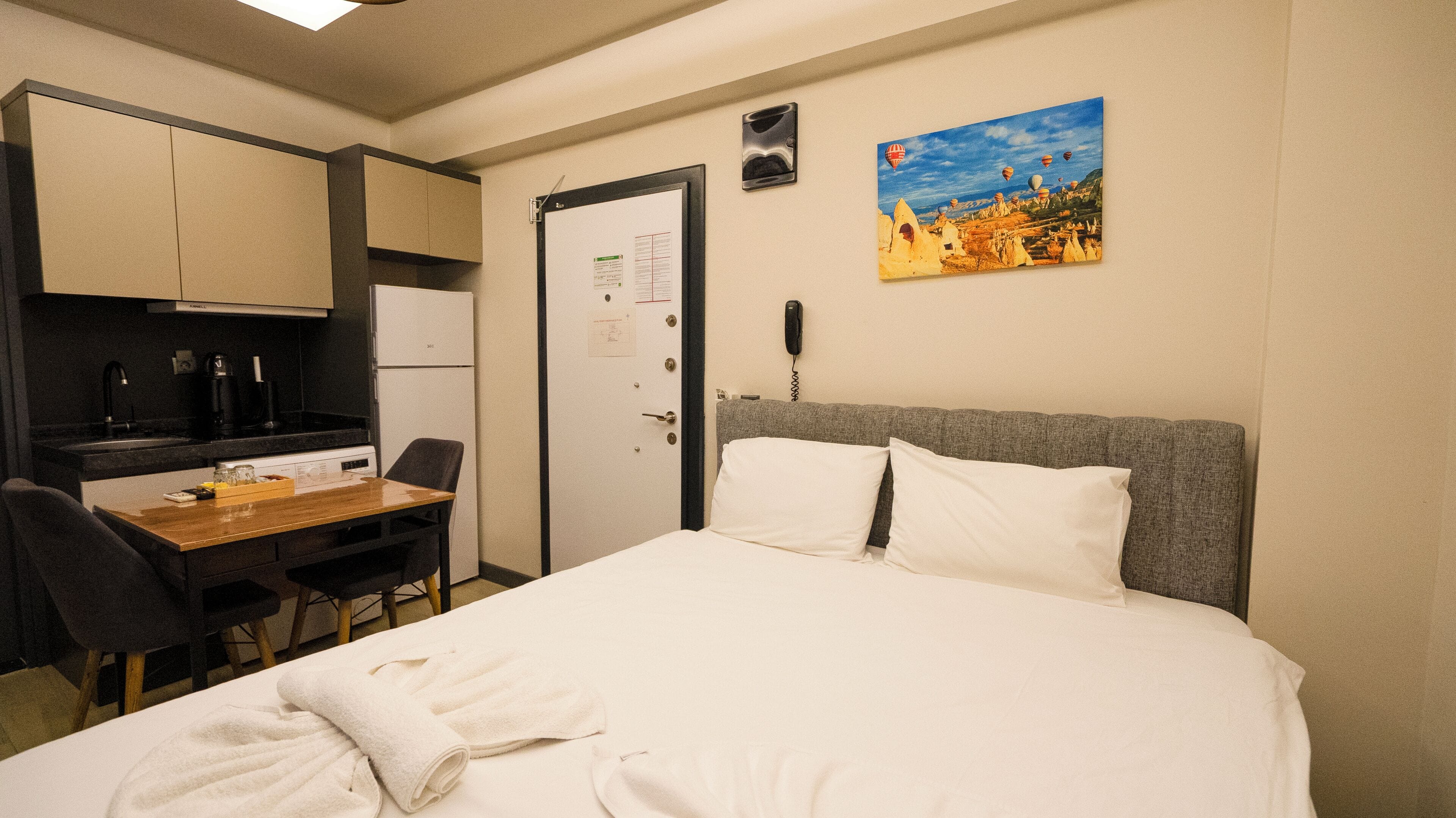 Quarto de hotel com cama, escrivaninha com cadeira, cozinha compacta e um quadro na parede.