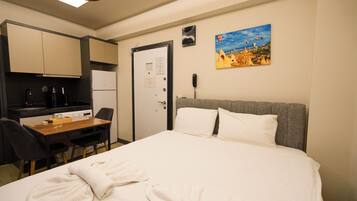 Quarto de hotel com cama, escrivaninha com cadeira, cozinha compacta e um quadro na parede.