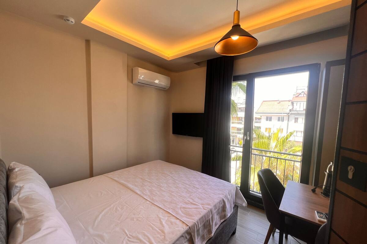 A hotel room with a bed, a desk, a chair, an air conditioning unit, a TV, and a balcony with a view of buildings and trees.