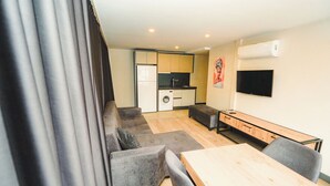 Een moderne woonkamer met een grijze bank, een houten eettafel en een keuken met een wasmachine en een flatscreen televisie.