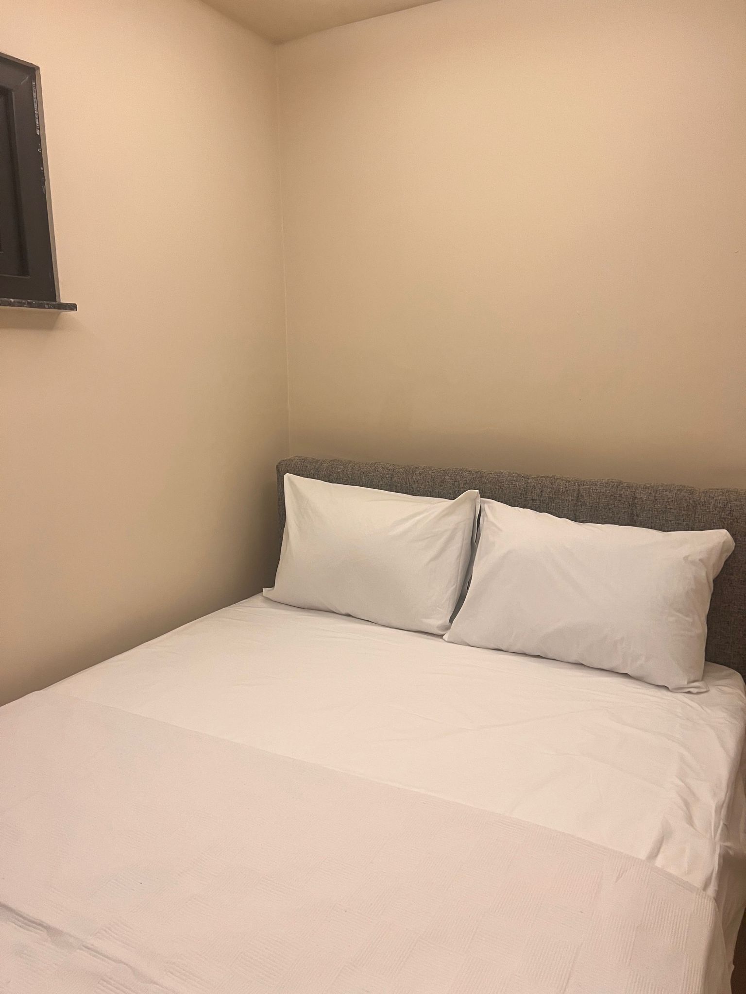 Una cama individual con ropa blanca y cabecera gris en una habitación con paredes color crema.