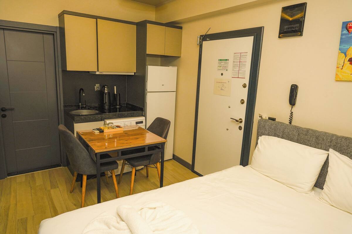 A compact hotel room with a bed, a small kitchenette, a dining table with chairs, and a door with a phone and a calendar.