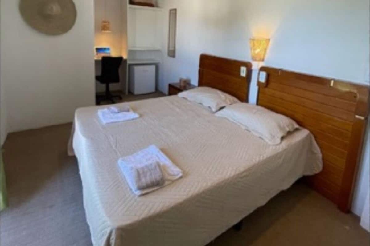 Cama bem arrumada com cabeceira de madeira, luminária de mesa e chapéu fixado na parede.