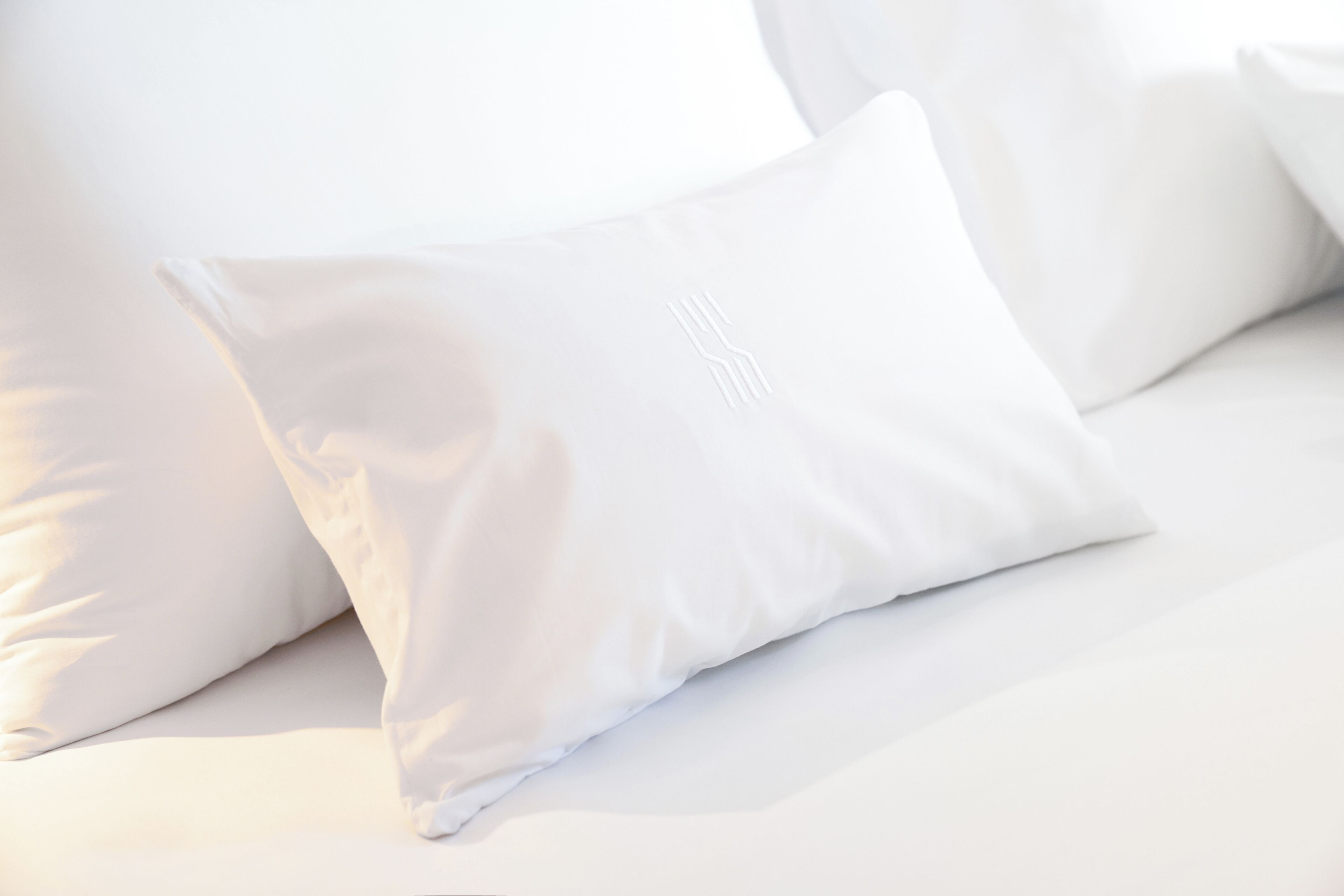 A bed with white bedding and pillows, a visible logo on one pillow, and a clean, minimalist design.
