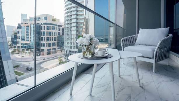 A modern balcony with white furniture, a vase of white flowers, and a view of city buildings. - Lusso Hometels J One (Dubai)