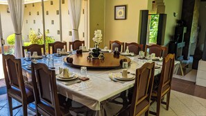 A dining table set for a meal with chairs, a vase of flowers, and a view of greenery outside. - Zambezi Family Lodge (Victoria Falls)