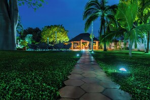 Un sendero conduce a un edificio con techo de paja, rodeado de palmeras y un jardín iluminado.