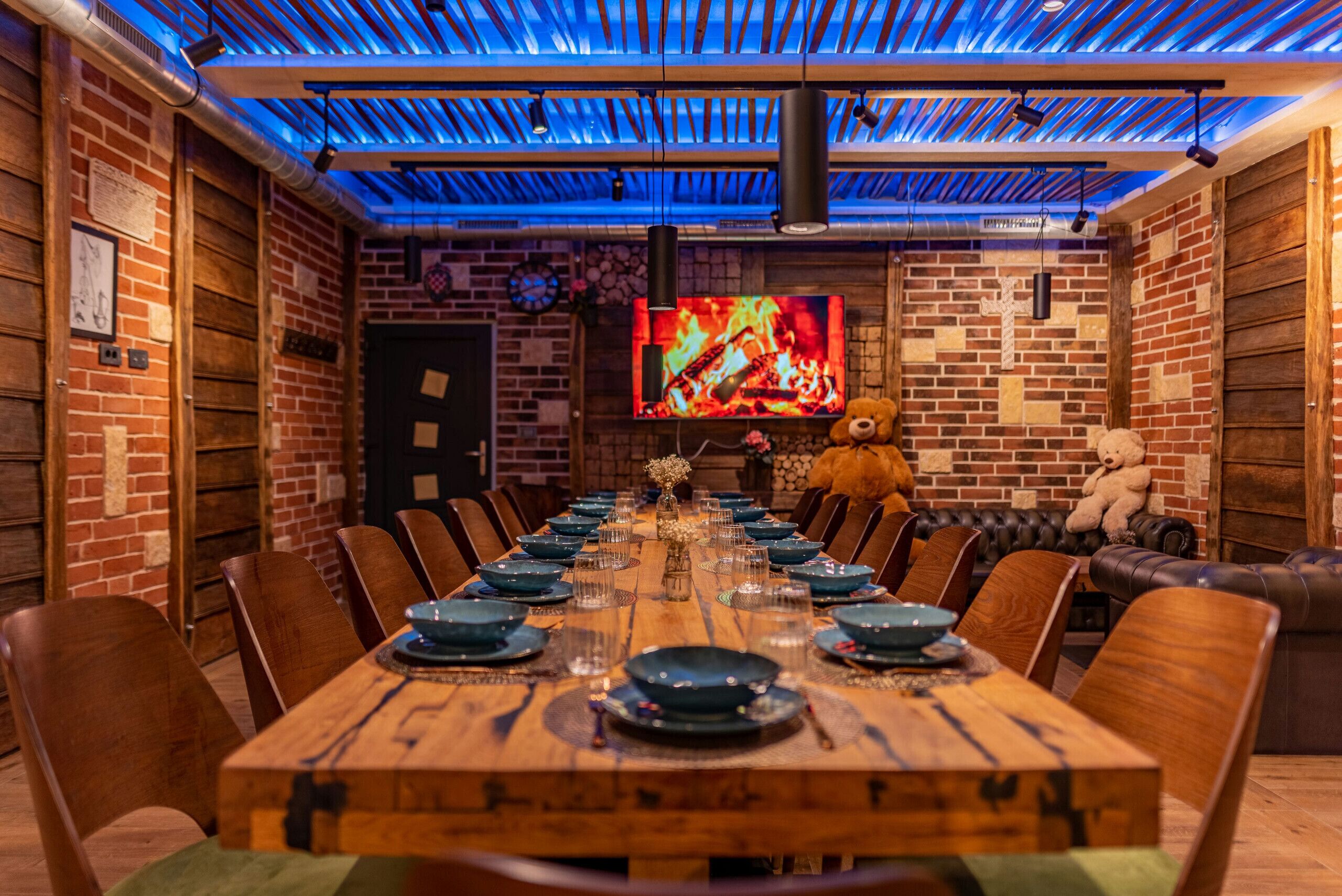 Une longue table à manger en bois, dressée avec des assiettes et des verres, entourée de fauteuils en cuir, dans une pièce aux murs de briques et dotée d’un grand écran de télévision diffusant une image de feu de cheminée.