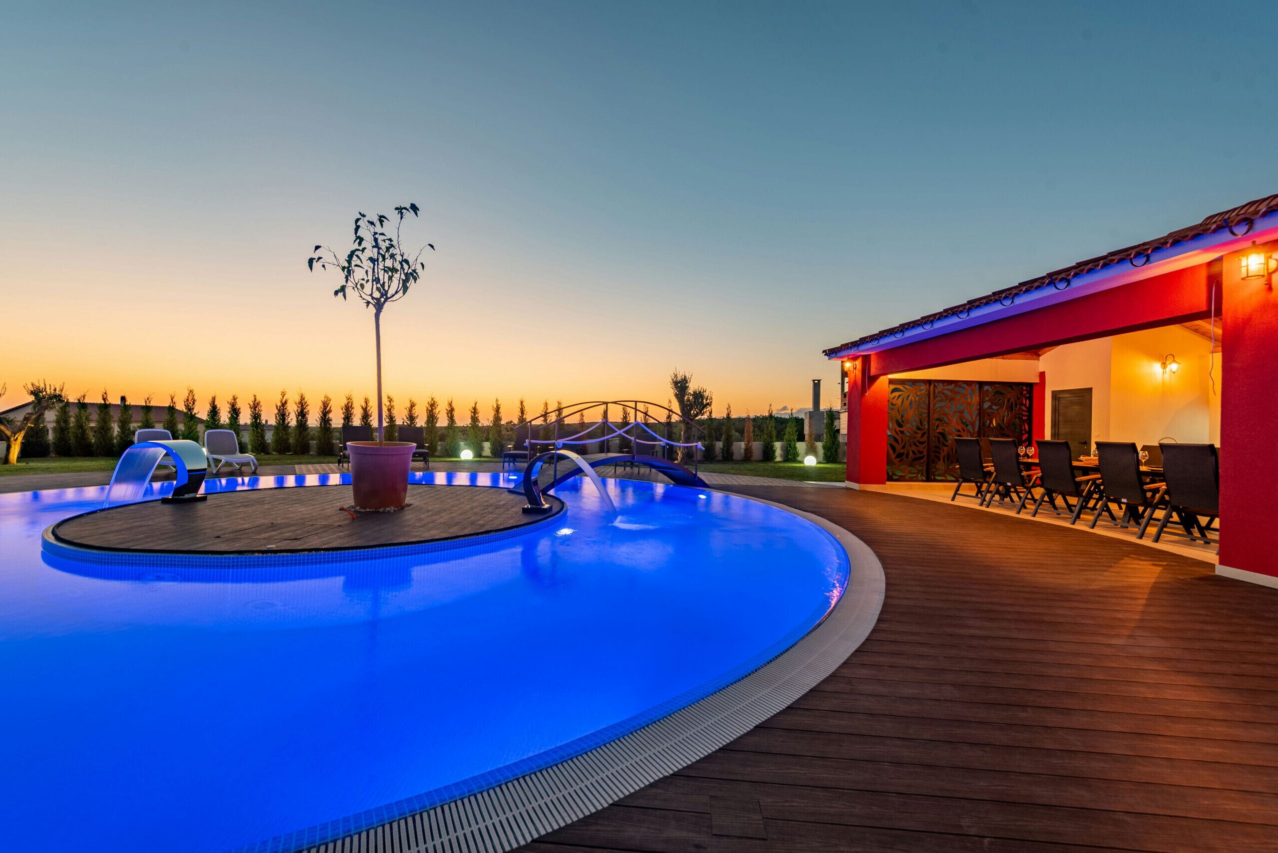 A swimming pool with a wooden deck, a covered seating area, and a view of the sunset.