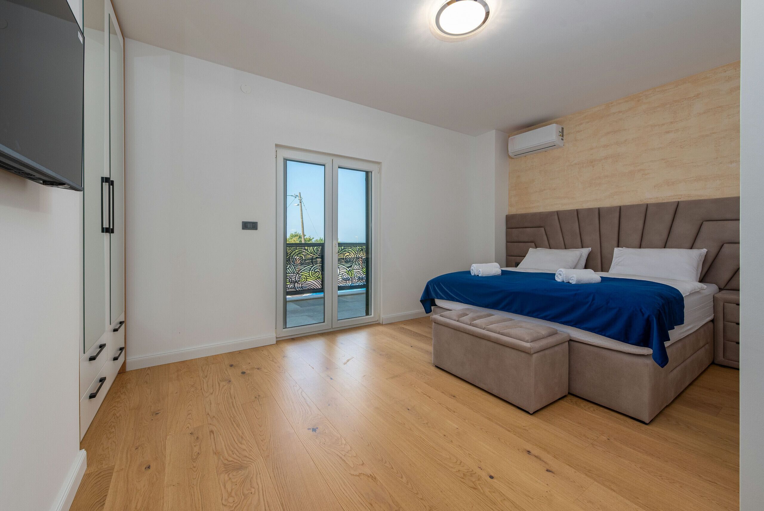 A bedroom with a bed, a sliding glass door to a balcony, a flat-screen TV, and a built-in air conditioning unit.