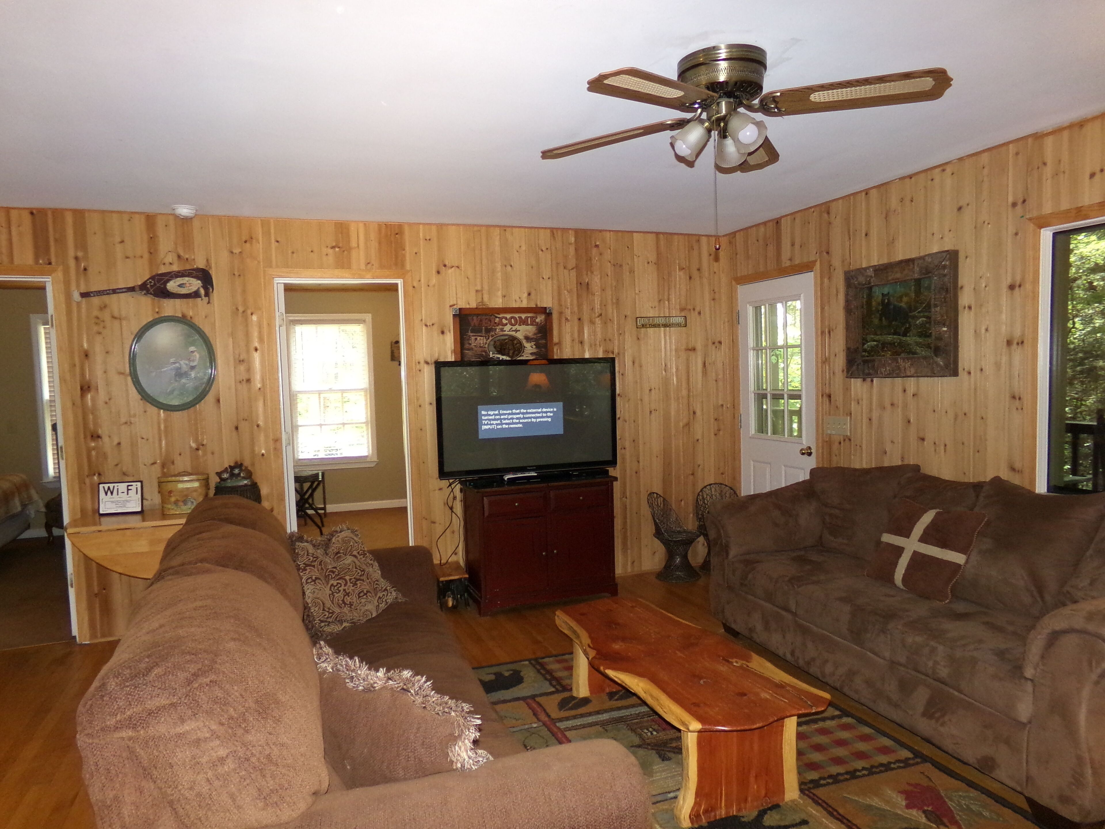 A wooden-paneled living room with a ceiling fan, a television, a sofa, a coffee table, and a door leading outside.