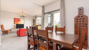 A modern dining area with a wooden table and chairs, a red sofa, a TV, and a city view through large windows. - Amber 2-Bedroom Apartment in Jal El Dib  (Jal El Dib)